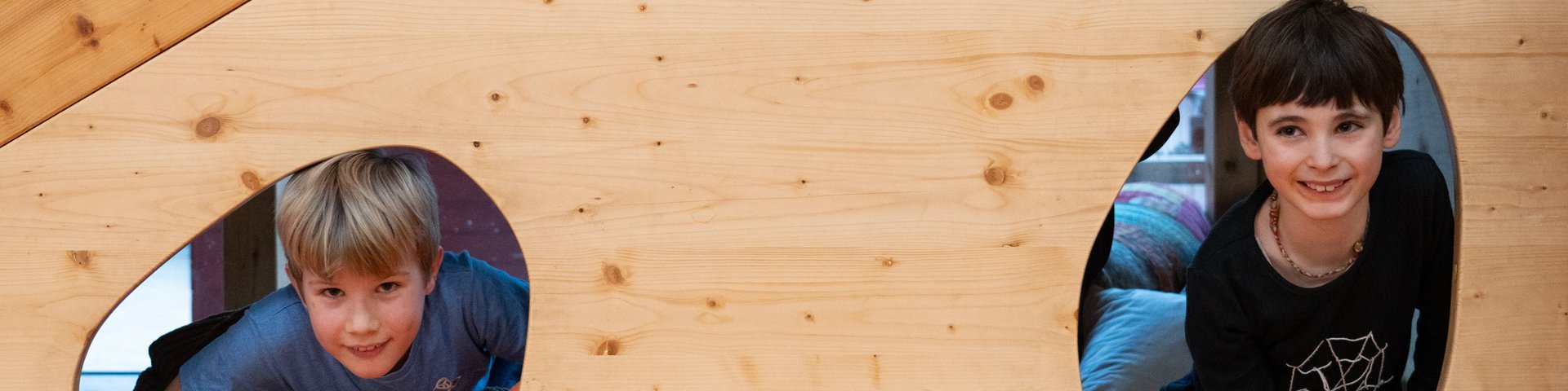 Drei Kinder lächeln und schauen durch unregelmäßig geformte Ausschnitte in einer großen Holztafel, von denen zwei unten und eines oben angebracht sind, und schaffen so eine spielerische und fröhliche Szene.