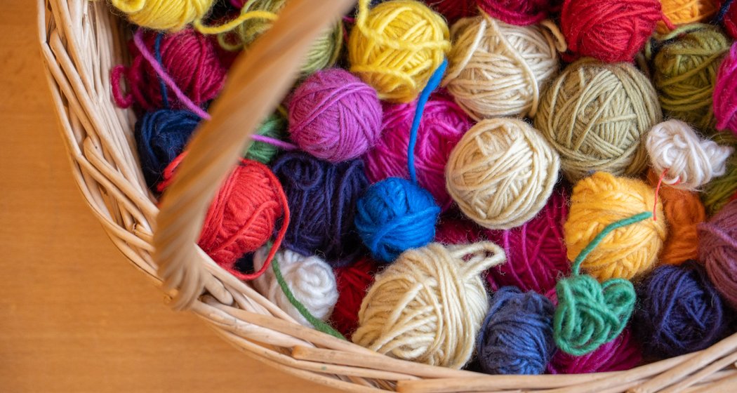 Ein Weidenkorb, der mit bunten Garnknäueln gefüllt ist, darunter rote, gelbe, blaue, grüne und beigefarbene, die auf einer Holzfläche liegen.