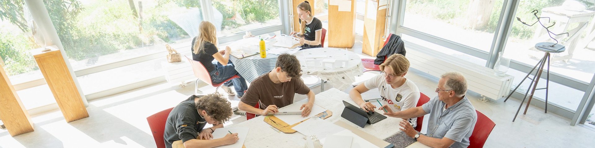 Fünf Personen sitzen um einen Tisch in einem hellen Raum mit großen Fenstern und arbeiten an Papieren und Modellen. Das Sonnenlicht strömt herein, und die Kunstmaterialien sind auf dem Tisch verstreut, während sie gemeinsam an einem Projekt arbeiten.