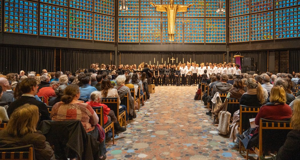 Ein Chor singt vor einem sitzenden Publikum in einer modernen Kirche mit blauen Glaswänden und einer großen goldenen Christusfigur über dem Altar.