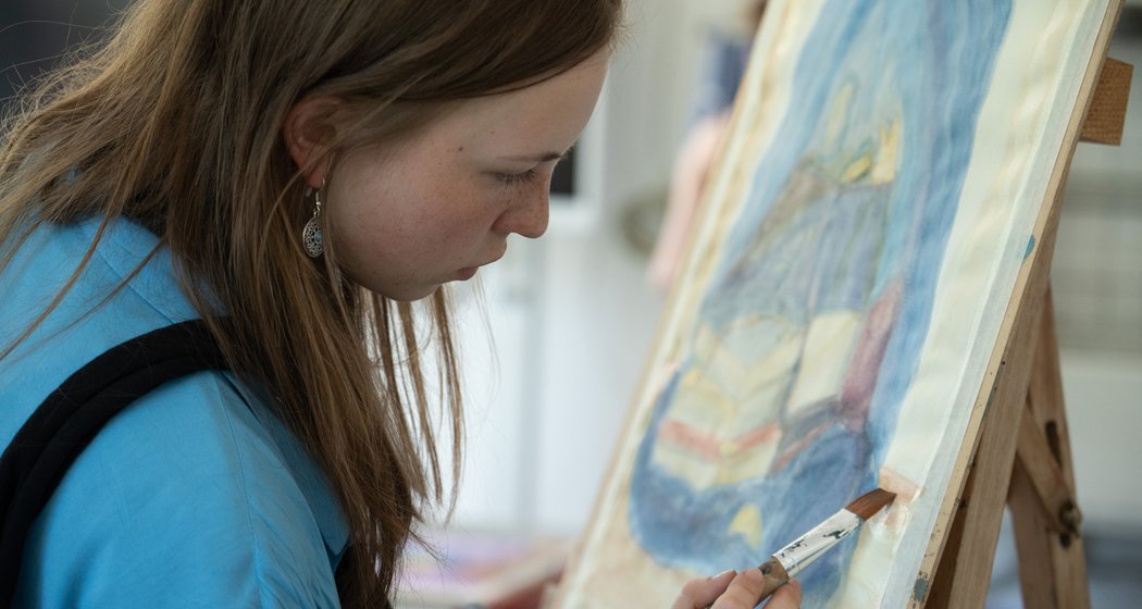 Eine junge Frau mit hellbraunem Haar malt konzentriert auf einer Leinwand, die auf einer Staffelei steht. Sie trägt ein blaues Hemd und hält einen Pinsel in der Hand, mit dem sie sorgfältig Details in ihr Kunstwerk einfügt.