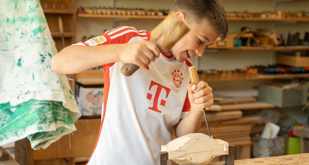 Ein lächelnder Junge in einem rot-weißen Sporthemd schnitzt ein Holzobjekt in einer Werkstatt voller Holzbearbeitungswerkzeuge und -materialien.